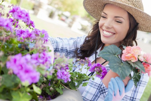 A woman wearing a wide-brimmed straw hat and gardening gloves is smiling as she tends to a collection of vibrant flowering plants in a well-maintained outdoor garden space. The garden features a lush lawn with dense, evenly cut grass, bordered by flower beds containing purple, pink, and orange blossoms. Behind her, there are hints of a paved patio area and a background of trees with green foliage, indicating a bright, sunny day. The scene suggests a typical residential garden in Walthamstow, with a tidy layout and healthy plant growth, reflecting professional garden care and landscaping maintenance. Lawn Mowing Walthamstowâ€™s gardening services are associated with ensuring lush lawns and vibrant plant displays in local outdoor spaces, aligning with the seen garden features.