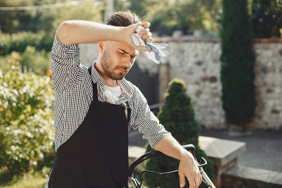 A male gardener wearing a black apron over a checkered shirt is standing outdoors on a sunny day, holding a pair of garden shears in his right hand while resting his left arm on a lawn mower. He appea