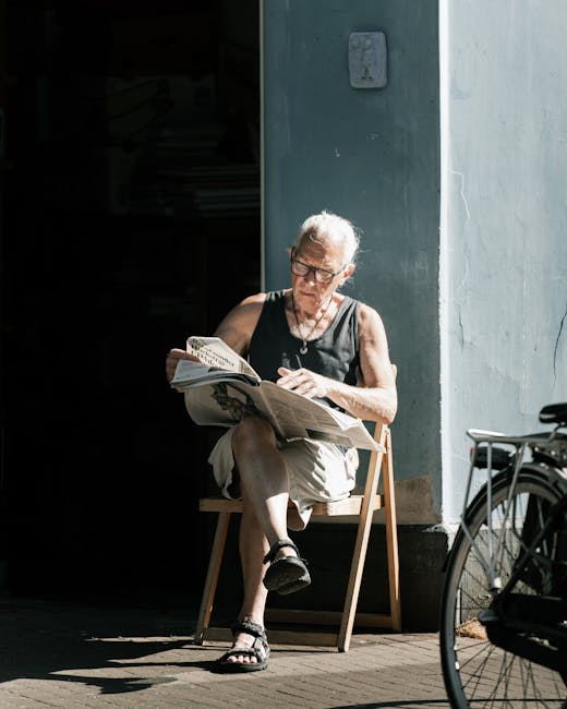 A male gardener wearing a black apron over a checkered shirt is standing outdoors on a sunny day, holding a pair of garden shears in his right hand while resting his left arm on a lawn mower. He appea