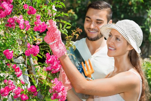 A man and woman in a lush garden with vibrant pink flowering bushes. The woman, wearing a white sun hat and gardening gloves, is carefully pruning or tending to the bushes with a small garden tool, while the man, dressed in a light-colored shirt, stands close by, smiling and observing. The garden features a well-maintained lawn, green foliage, and a backdrop of trees and shrubbery, indicating a cultivated outdoor space in Walthamstow. The scene suggests active gardening work, supported by local gardening services that enhance outdoor areas through lawn mowing, pruning, and landscape maintenance. The natural daylight highlights the rich colours of the flowers and greenery, creating a lively and inviting outdoor environment, typical of a landscaped Walthamstow garden managed by Lawn Mowing Walthamstow or similar gardening specialists.